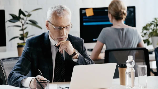 Empresario en gafas que escribe en el cuaderno y mirando el ordenador portátil cerca de colega borrosa en la oficina - foto de stock