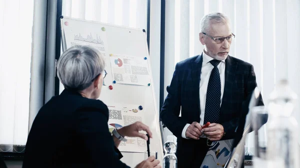 Homme d'affaires mature debout près de flip chart et collègue flou dans le bureau — Photo de stock