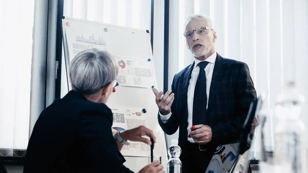 Uomo d'affari in abbigliamento formale parlando vicino flip chart e collega in ufficio — Foto stock