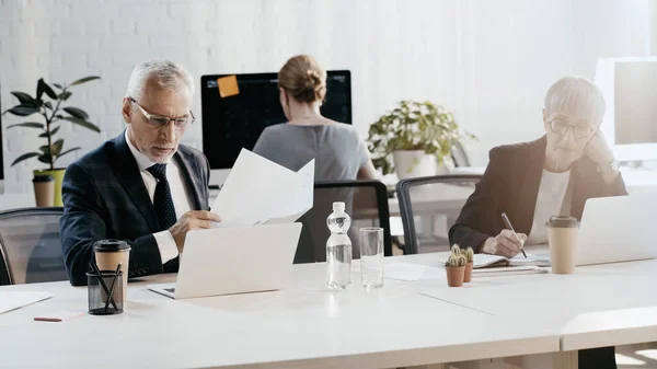 Homme d'affaires mature tenant des documents près d'un ordinateur portable et collègue travaillant dans le bureau — Photo de stock