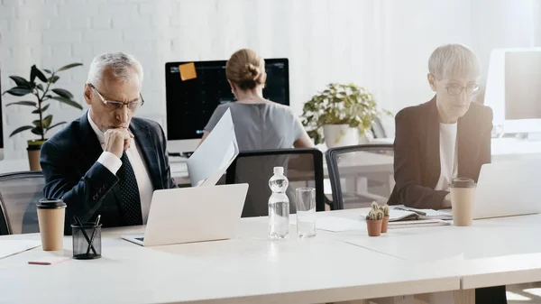 Empresario sosteniendo papeles cerca de la computadora portátil y colega trabajando en la oficina - foto de stock