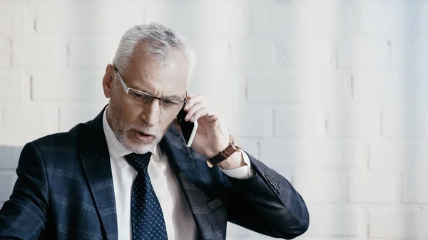 Mature businessman in eyeglasses talking on smartphone in office — Stock Photo