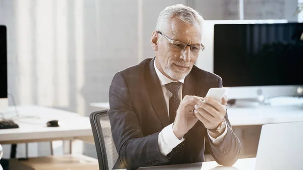 Lächelnder Geschäftsmann mit Handy in der Nähe von Laptop im Büro — Stockfoto