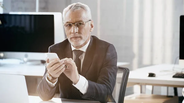 Homme d'affaires d'âge moyen utilisant un téléphone portable et regardant la caméra dans le bureau — Photo de stock