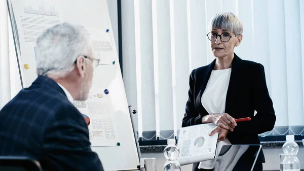 Middle aged businesswoman holding clipboard near colleague and flip chart in office — Stock Photo