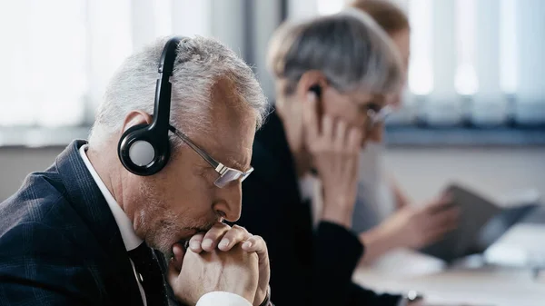 Vista lateral del pensativo hombre de negocios con auriculares sentado cerca de colegas borrosos en la oficina - foto de stock