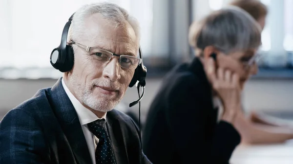 Homme d'affaires mature dans un casque regardant la caméra près de collègues flous au bureau — Photo de stock