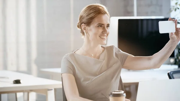 Позитивная деловая женщина делает селфи на смартфоне рядом с ноутбуком в офисе — стоковое фото