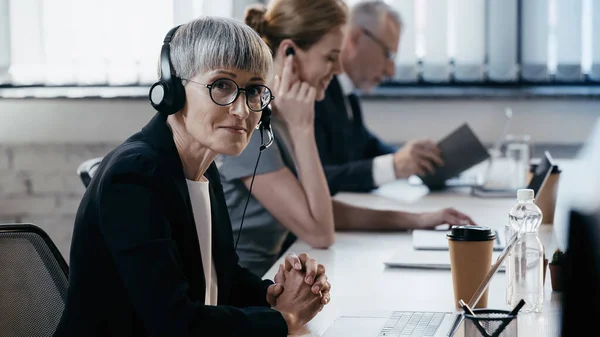 Madura mujer de negocios en auriculares mirando la cámara cerca de la computadora portátil y café para ir en la oficina - foto de stock