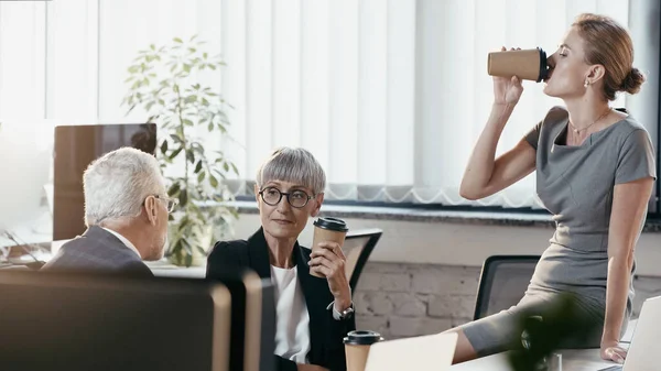 Businesswoman drinking coffee to go near mature colleagues in office — Stock Photo