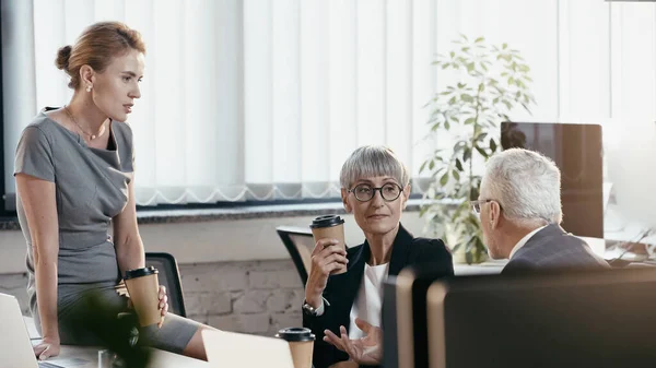 Les gens d'affaires avec des tasses en papier parlant près des ordinateurs portables dans le bureau — Photo de stock