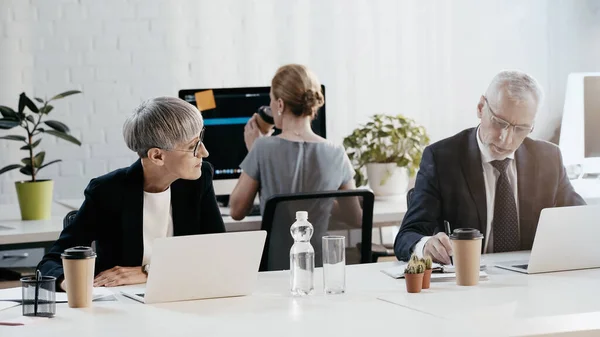 Businessman in formal wear writing on notebook near devices and colleagues in office — Stock Photo