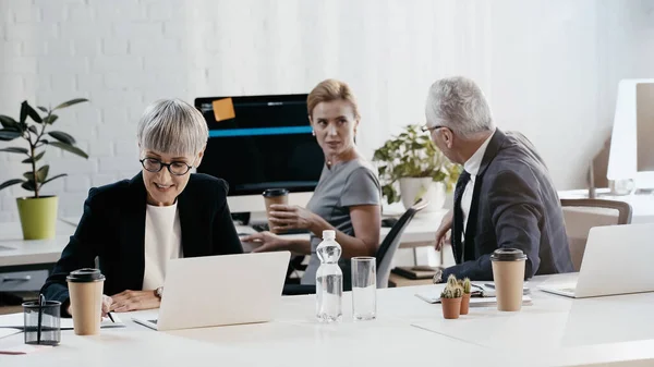 Lächelnde reife Geschäftsfrau schreibt auf Notizbuch, während Kollegen im Büro reden — Stockfoto