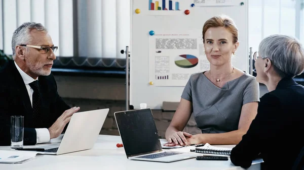 Positive Geschäftsfrau blickt in die Kamera neben Kollegen mittleren Alters und Flipchart im Büro — Stockfoto