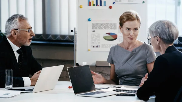 Бізнес-леді дивиться на зрілого колегу біля пристроїв під час зустрічі в офісі — стокове фото
