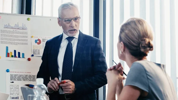 Uomo d'affari maturo che parla con collega vicino a grafici su lavagna a fogli mobili in ufficio — Foto stock