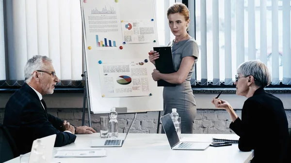 Business people in formal wear talking near devices and flip chart in office — Stock Photo