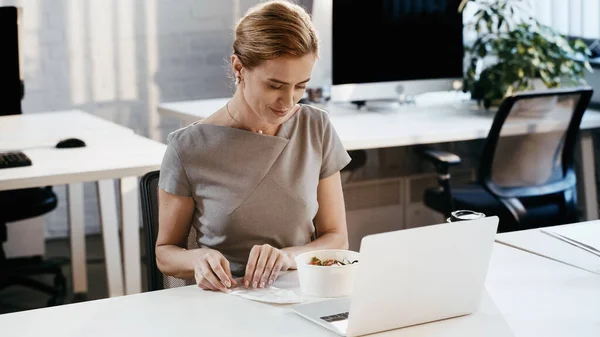 Femme d'affaires à la recherche de salade à emporter près d'un ordinateur portable au bureau — Photo de stock