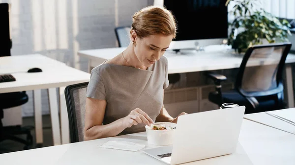 Gestionnaire tenant fourchette près de salade à emporter et ordinateur portable dans le bureau — Photo de stock