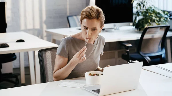 Empresaria comiendo ensalada fresca cerca de café para llevar y portátil en la oficina - foto de stock