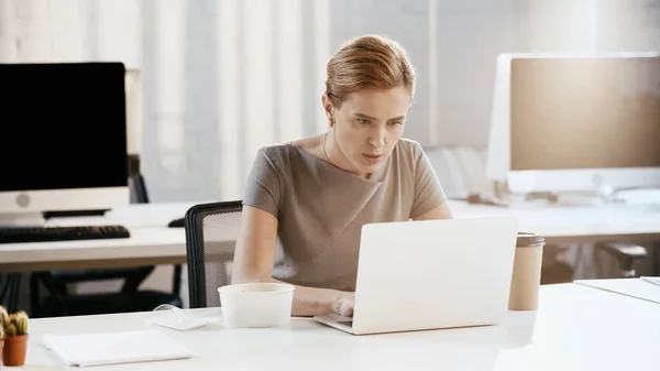Geschäftsfrau benutzt Laptop in der Nähe von Kaffee zum Mitnehmen und Container im Büro — Stockfoto
