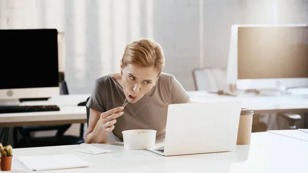 Donna d'affari che mangia insalata fresca vicino a computer portatile e carte in ufficio — Foto stock