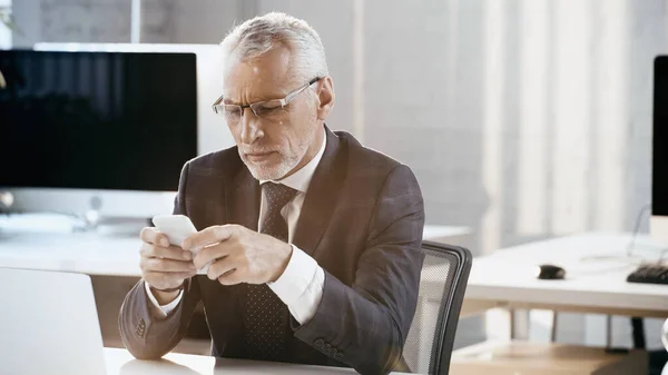 Mature businessman in eyeglasses using mobile phone near laptop in office — Stock Photo