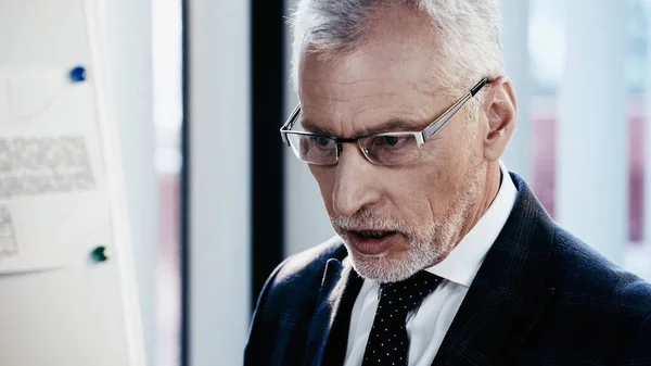 Middle aged businessman in eyeglasses standing near blurred flip chart in office — Stock Photo