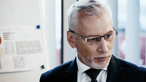 Middle aged businessman standing near blurred flip chart in office — Stock Photo