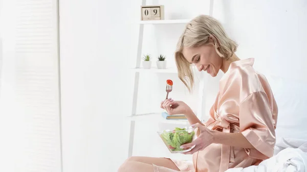 Веселая беременная женщина держит миску с салатом и смотрит на живот в спальне — стоковое фото