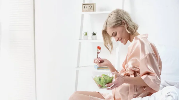 Cheerful pregnant woman holding bowl with salad and looking at belly in bedroom — Stock Photo