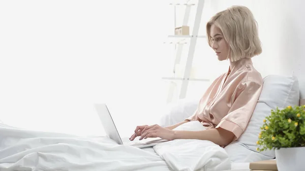 Freelance biondo usando il computer portatile mentre si siede a letto — Foto stock