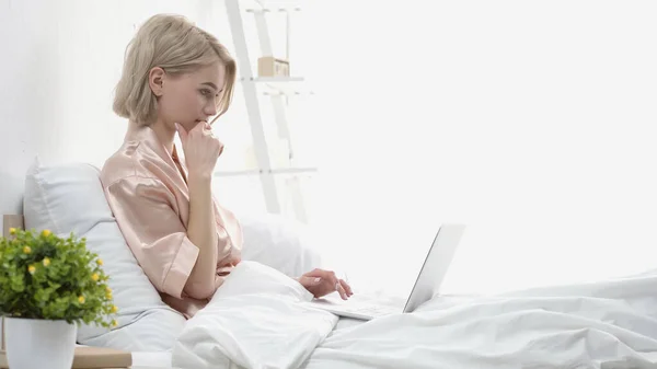 Задумчивая блондинка с помощью ноутбука, сидя в постели — стоковое фото