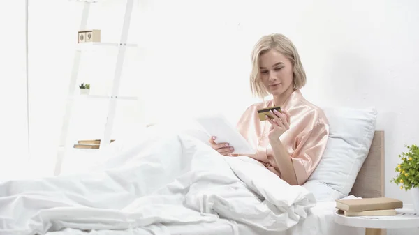 Blonde woman holding digital tablet and credit card while doing online shopping in bed — Stock Photo