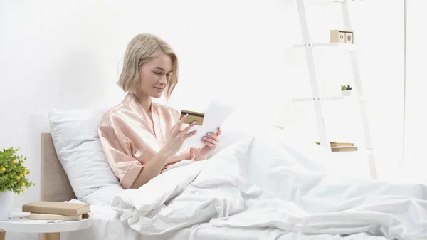 Donna sorridente che tiene tablet digitale e carta di credito mentre fa shopping online a letto — Foto stock