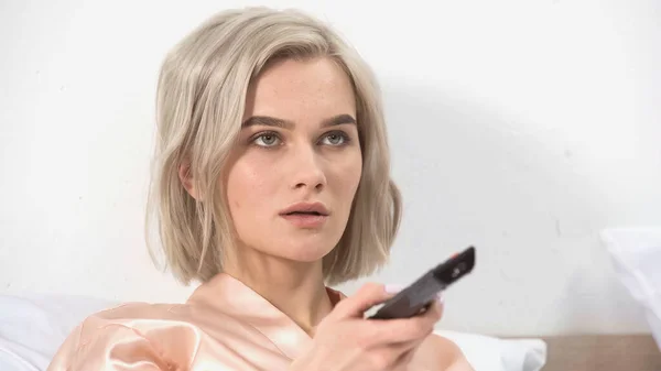 Shocked blonde woman holding remote controller in bedroom — Stock Photo