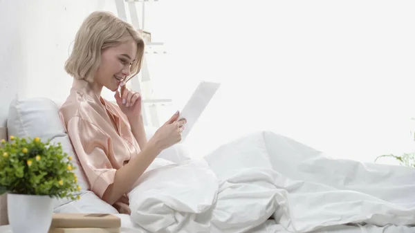 Vista lateral de mujer rubia feliz usando tableta digital y sonriendo en la cama - foto de stock