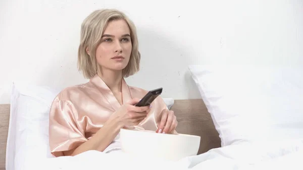 Blonde woman sitting with bowl while holding remote controller and watching movie — Stock Photo