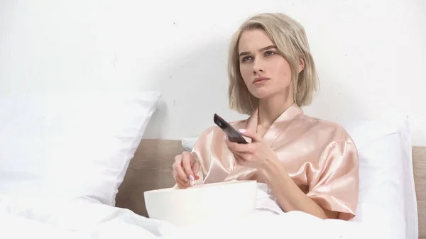 Giovane donna bionda seduta con ciotola mentre tiene il telecomando e guarda film a letto — Foto stock