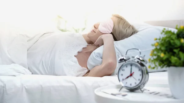 Jeune femme blonde dans le masque de sommeil couché dans le lit, étirant et souriant dans la chambre — Photo de stock