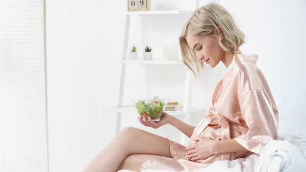 Vue latérale de la femme enceinte heureuse tenant bol avec salade et regardant le ventre dans la chambre — Photo de stock