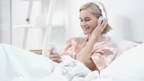 Счастливая молодая женщина в беспроводных наушниках слушать музыку и использовать смартфон — стоковое фото