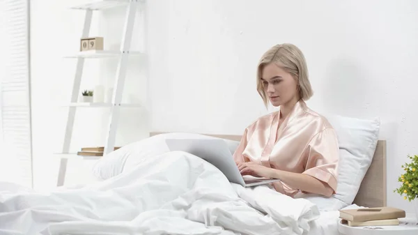 Jovem freelancer loira usando laptop enquanto sentado na cama — Fotografia de Stock