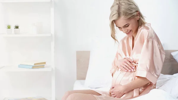 Cheerful pregnant woman touching belly and smiling while sitting in bedroom — Stock Photo