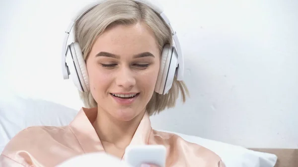 Cheerful young woman in wireless headphones listening music and using smartphone — Stock Photo