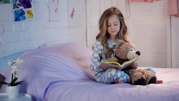 Preteen kid holding soft toy and reading book on bed — стоковое фото