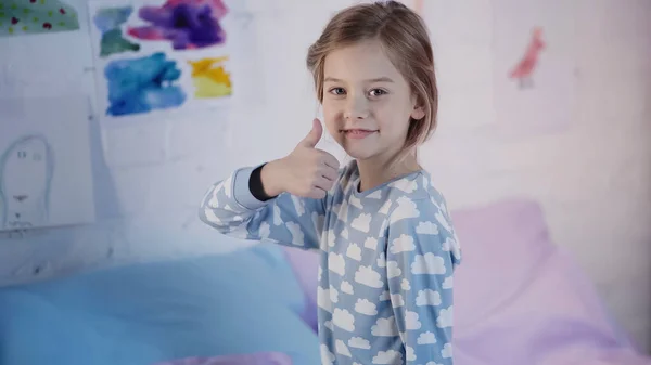 Smiling preteen child in pajama showing like gesture on bed at home — стоковое фото
