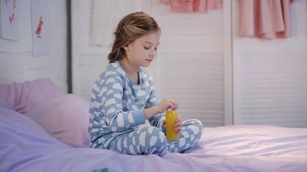 Preteen child in pajama holding bottle with orange juice on bed — Stockfoto