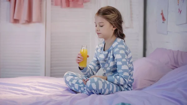 Girl in pajama holding bottle of orange juice on bed in evening — Stock Photo