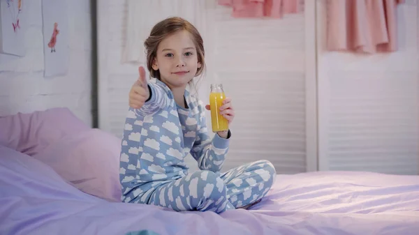 Smiling child in pajama holding orange juice and showing like gesture on bed — Stock Photo